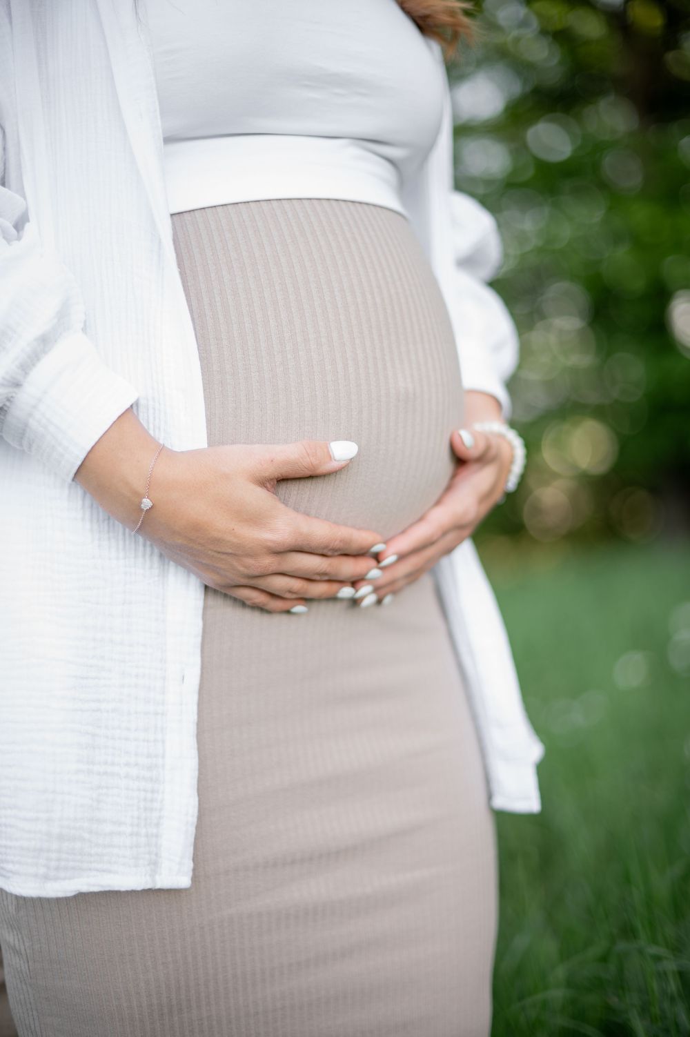 Nahaufnahme eines schwangeren Bauches in einem taillierten beigen Kleid mit Händen, die sanft den Bauch streicheln.