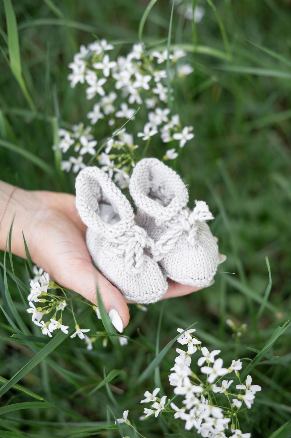 Eine Hand hält winzige gestrickte weiße Babyschühchen vor einem Hintergrund aus zarten weißen Wildblumen und grünem Gras.