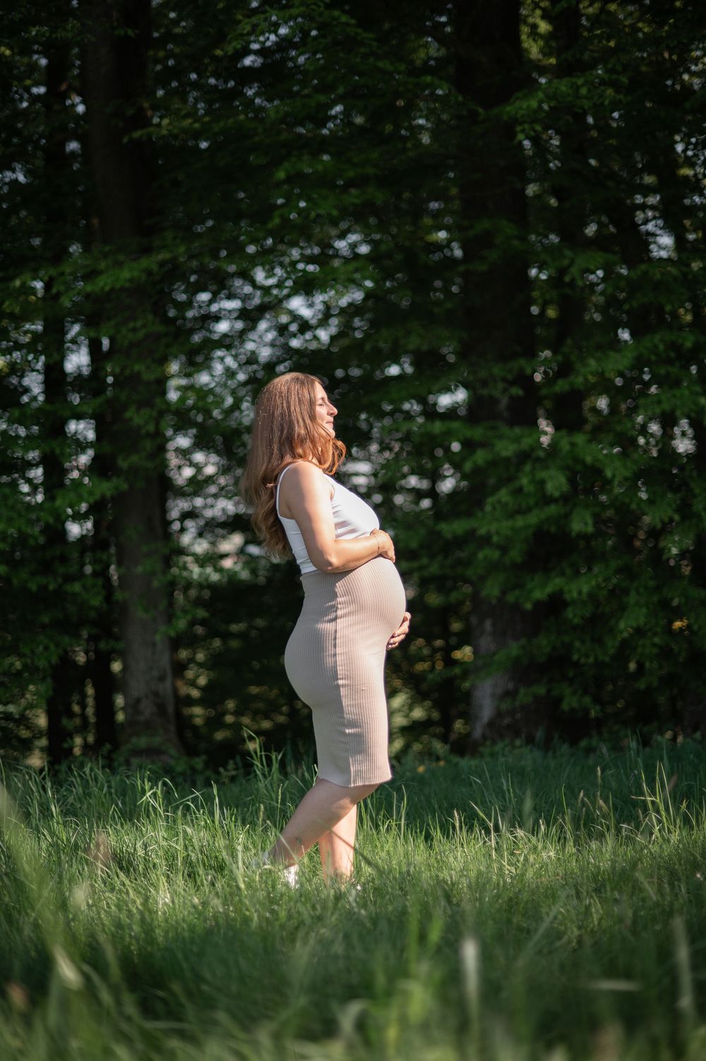 Eine schwangere Frau steht gelassen im hohen Gras und trägt ein tailliertes beiges Kleid vor einem Hintergrund aus üppigen Waldbäumen.