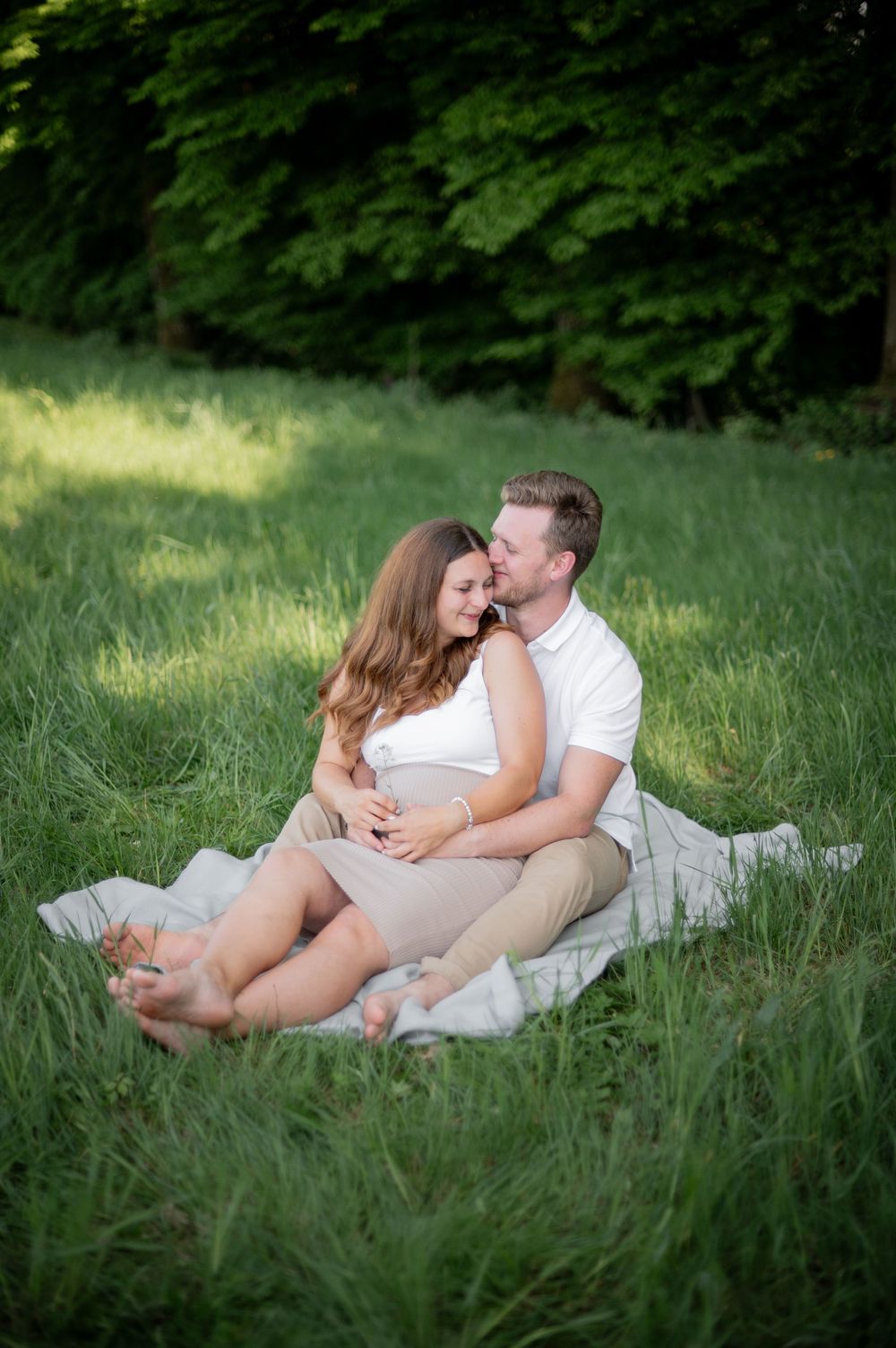 Ein romantisches Babybauchfoto zeigt ein Paar, das zusammen auf einer Decke im hohen Gras mit grünen Bäumen im Hintergrund sitzt.