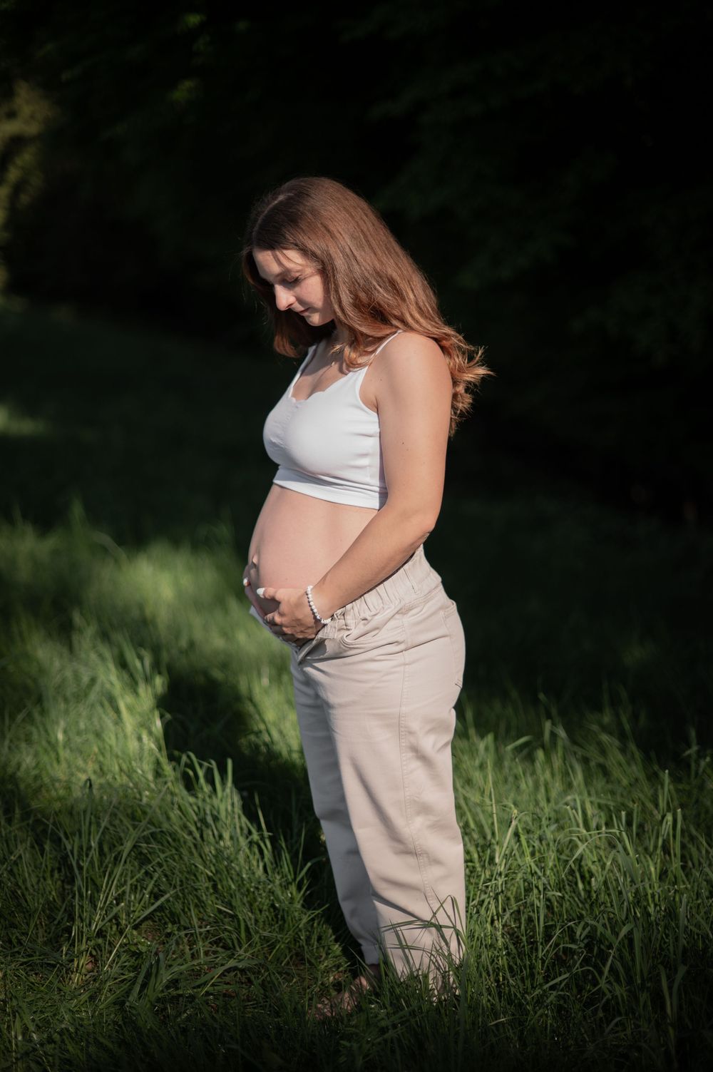 Wunderschöne Mutterschaftsfotos im Freien mit einem weißen Crop-Top und einer beigen Hose in einer natürlichen, grasbewachsenen Umgebung.