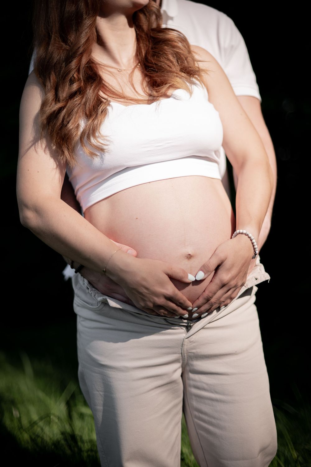 Nahaufnahme eines Schwangerschaftsfotos, das einen schwangeren Bauch mit Händen in Herzform zeigt. Sie trägt ein weißes Crop-Top und eine be