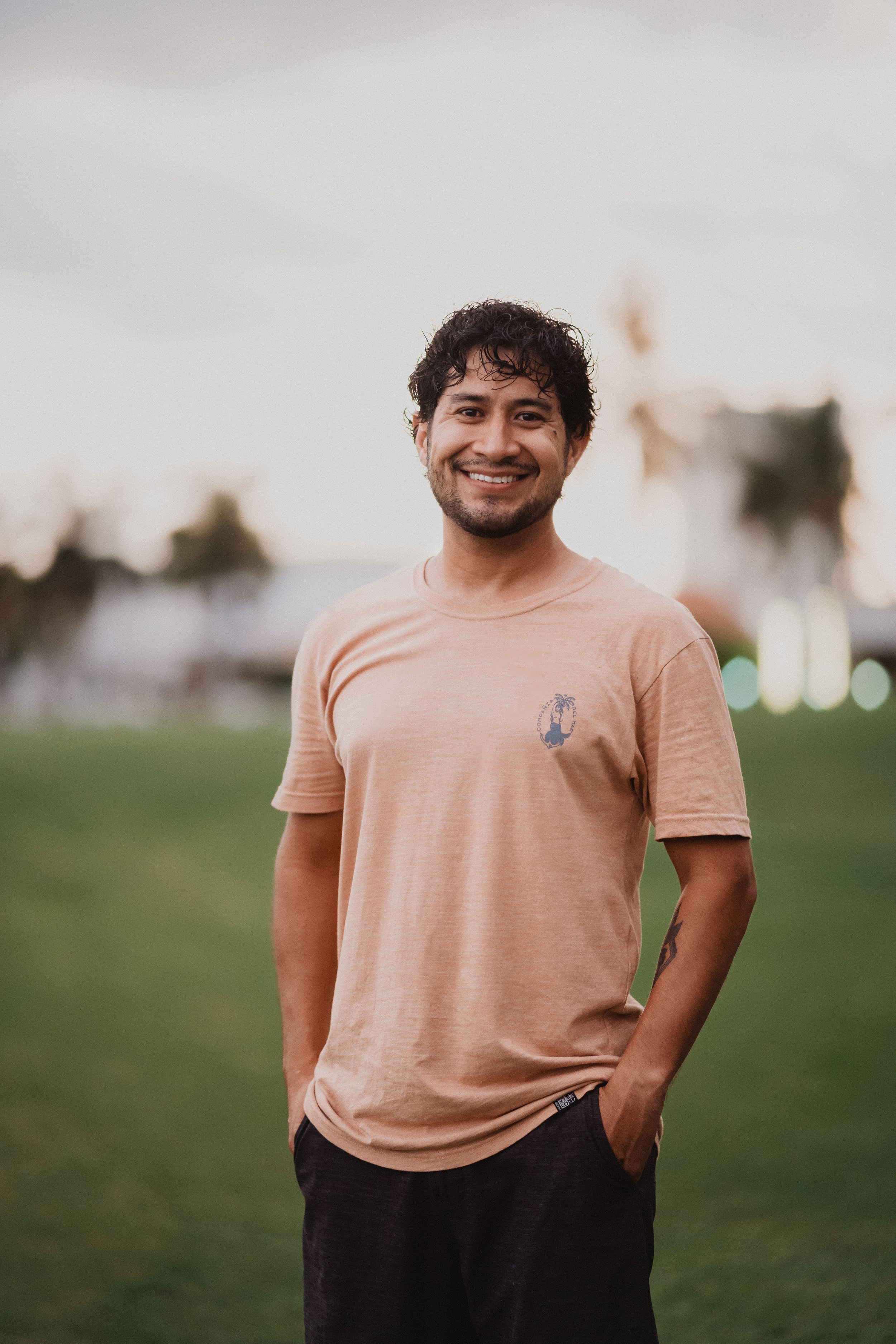 Person wearing a peach colored t-shirt with small anchor graphic stands smiling on grassy field with palm trees.