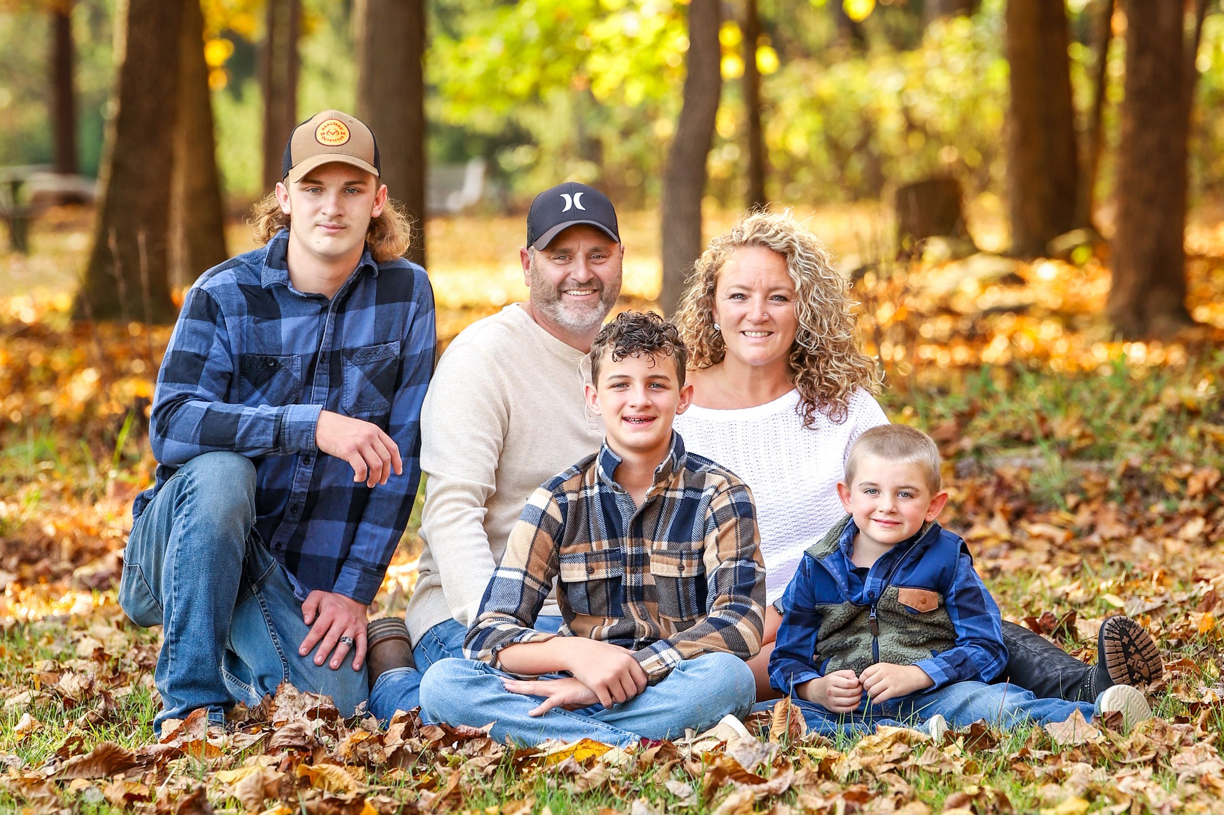 A family sits together on fallen autumn leaves in an outdoor setting surrounded by trees during fall.