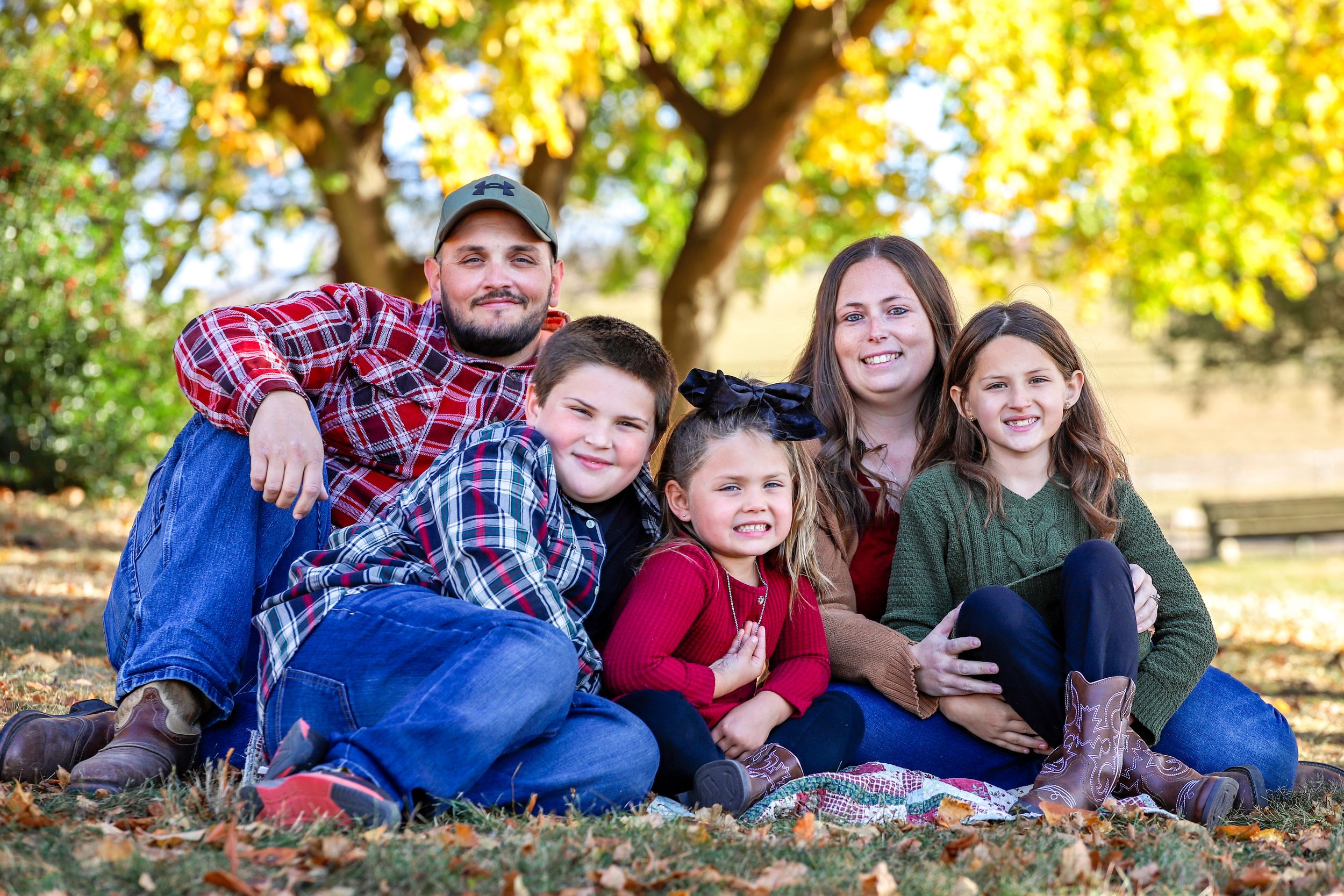 Family of five sitting together outdoors in autumn leaves wearing casual clothing and plaid shirts.