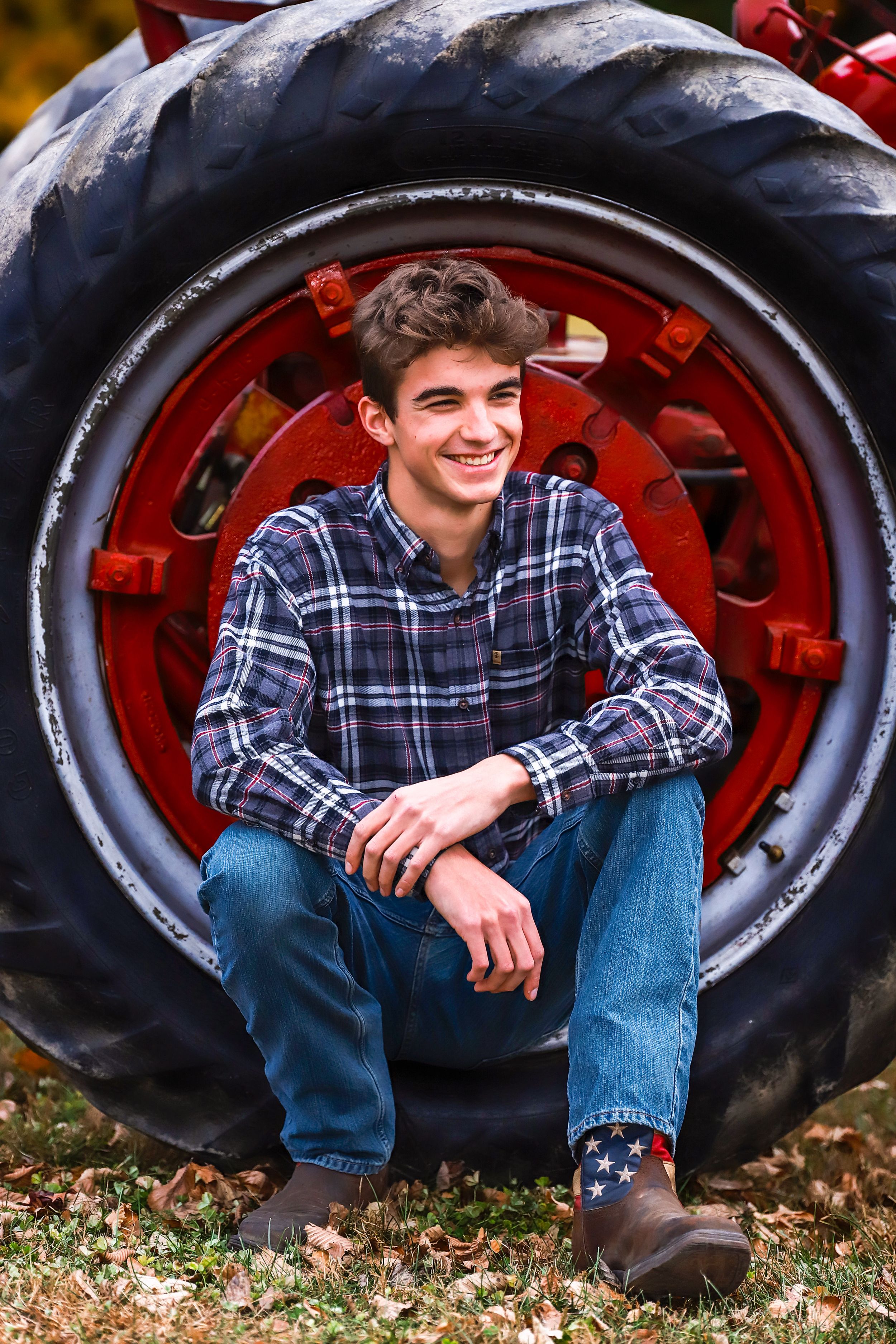 Someone sits casually in front of a large red tractor tire wearing a plaid flannel shirt and jeans in an autumn setting.
