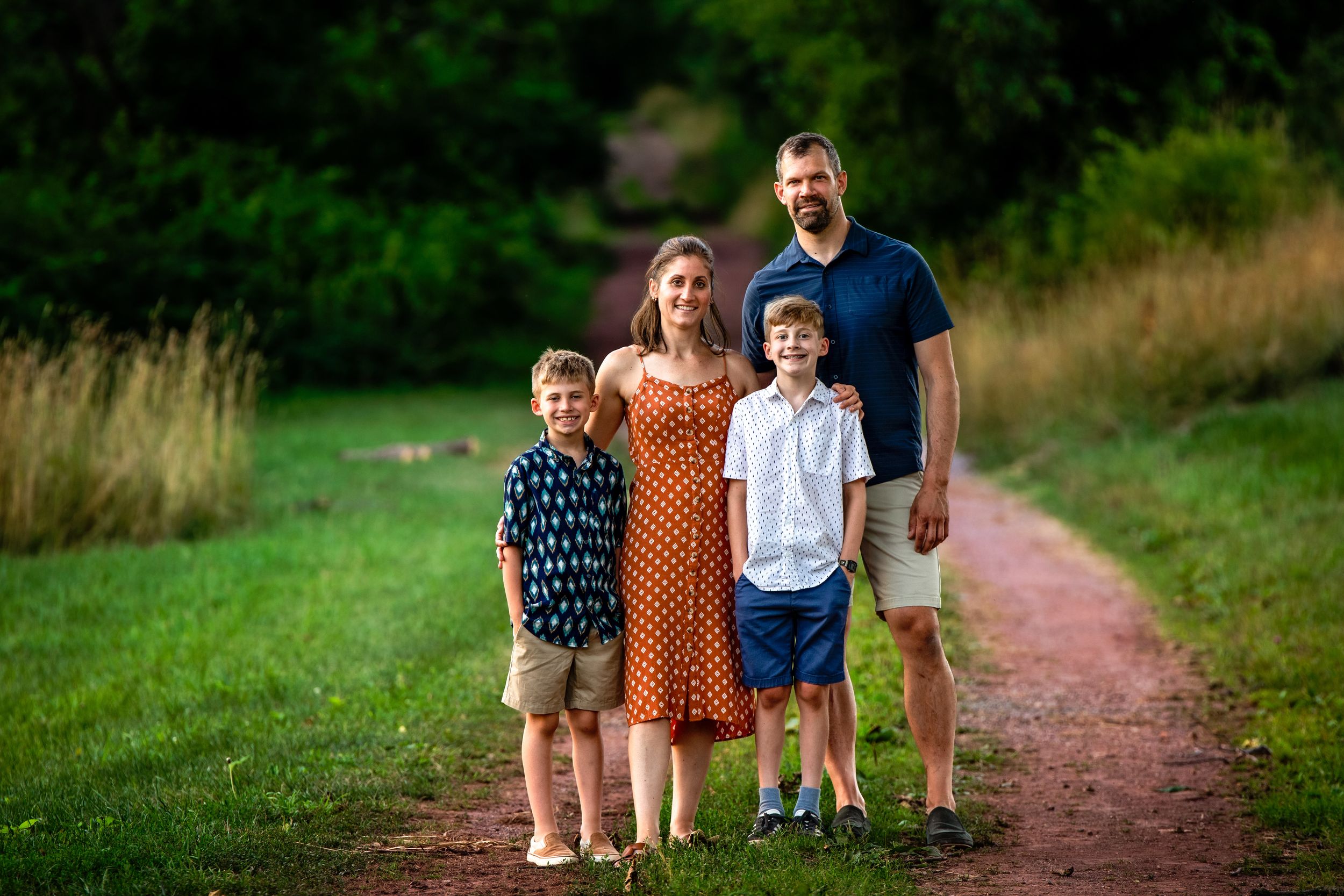 A family of four walks together on a peaceful dirt path surrounded by lush greenery on a summer day.