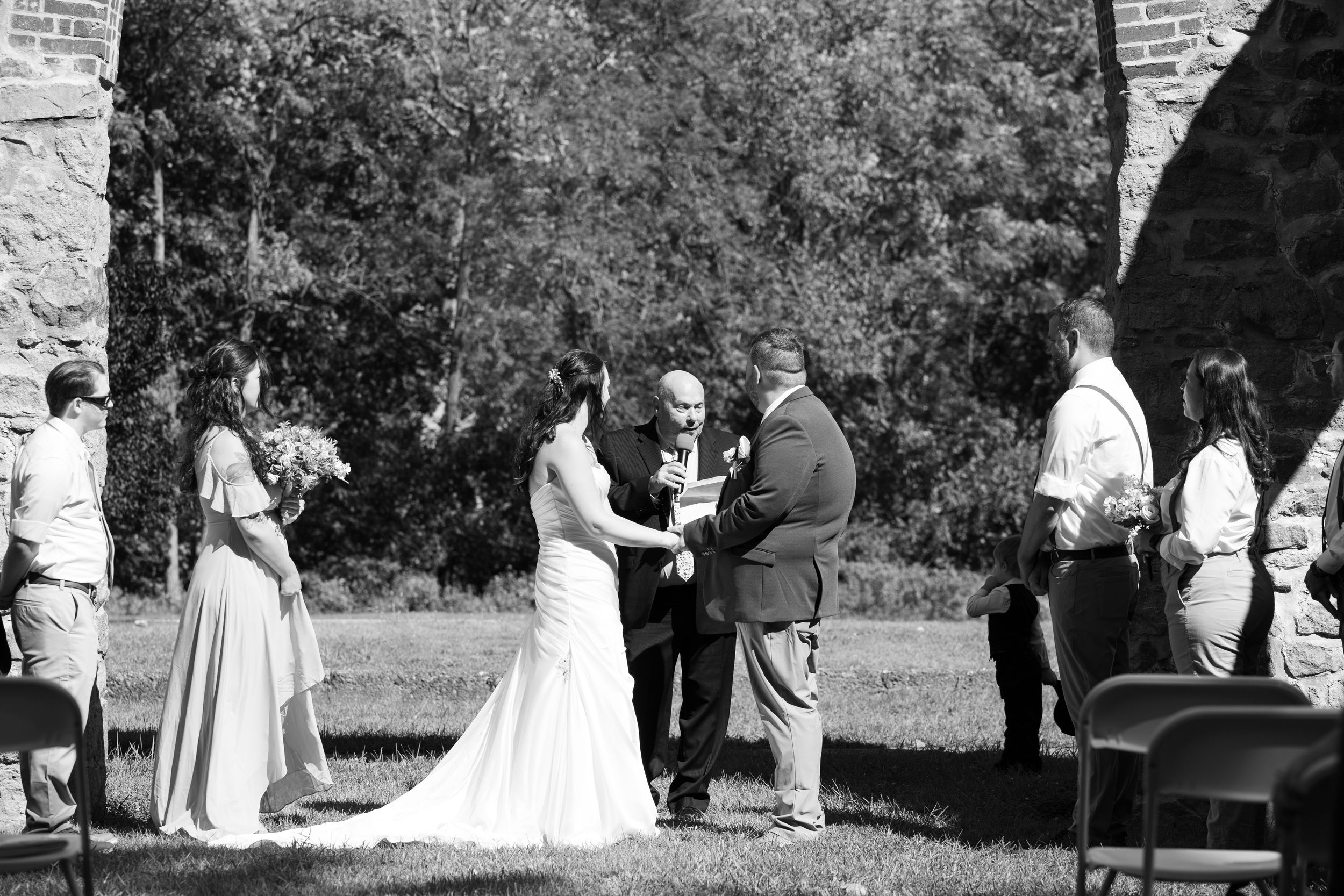 A black and white outdoor wedding ceremony taking place under stone archways with guests gathered around.