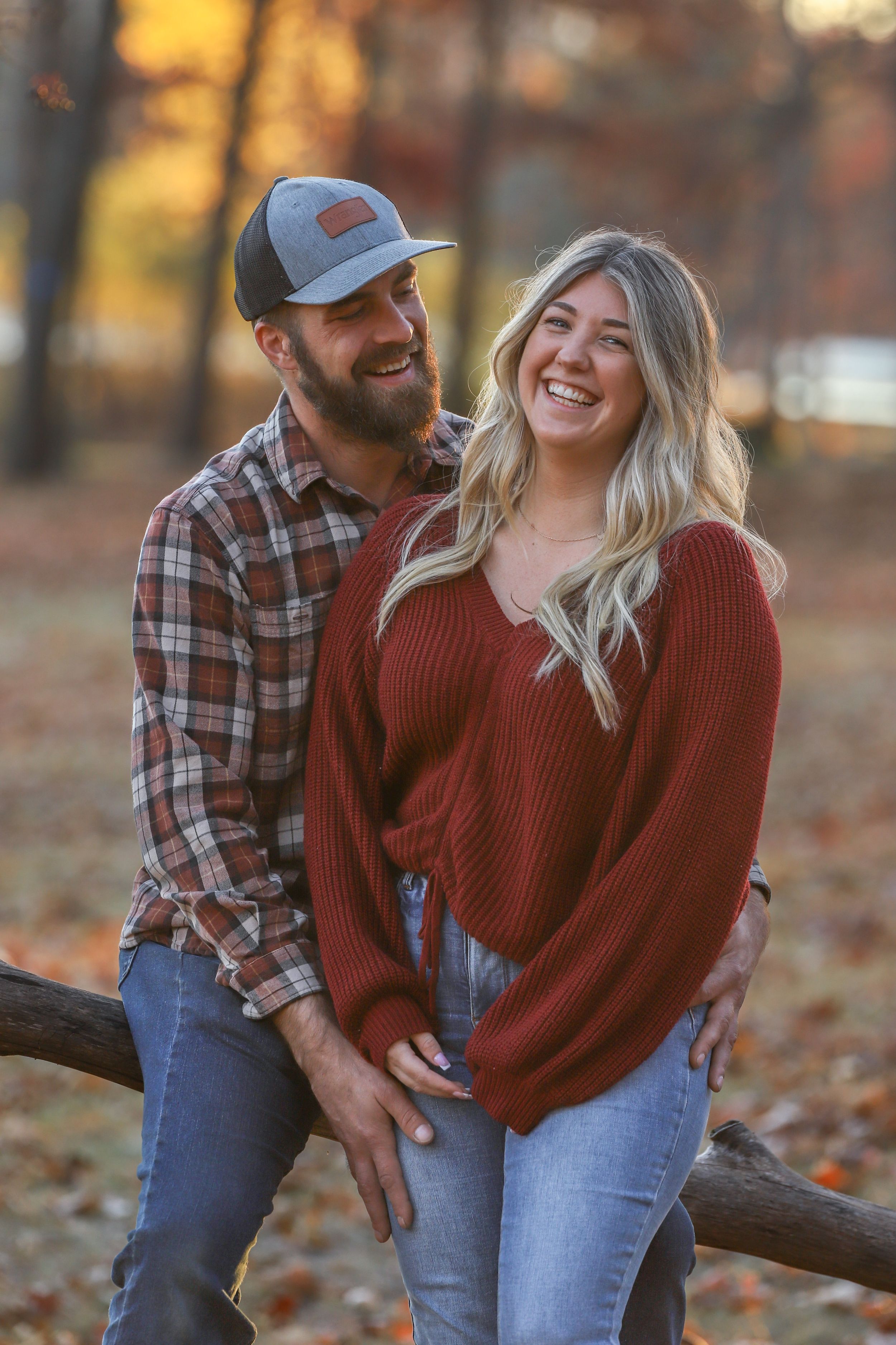 A couple in casual fall attire share a candid moment while leaning against a fence in an autumn forest setting.