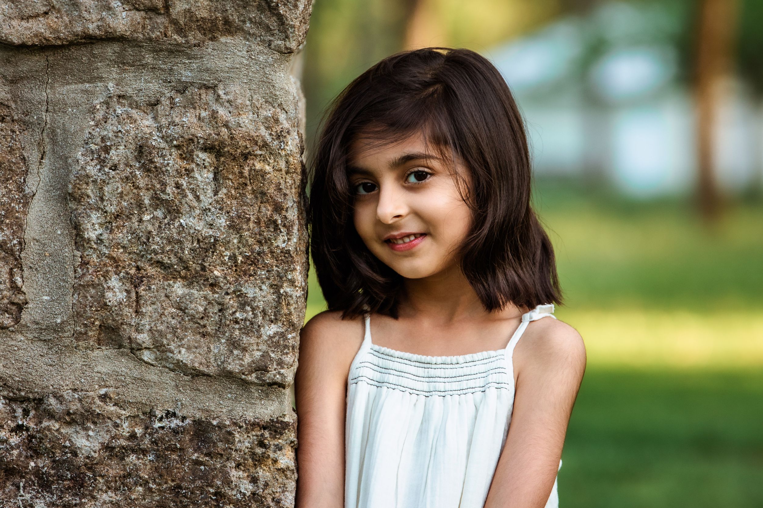 A young girl in a white summer dress stands near a stone wall outdoors with a soft smile.