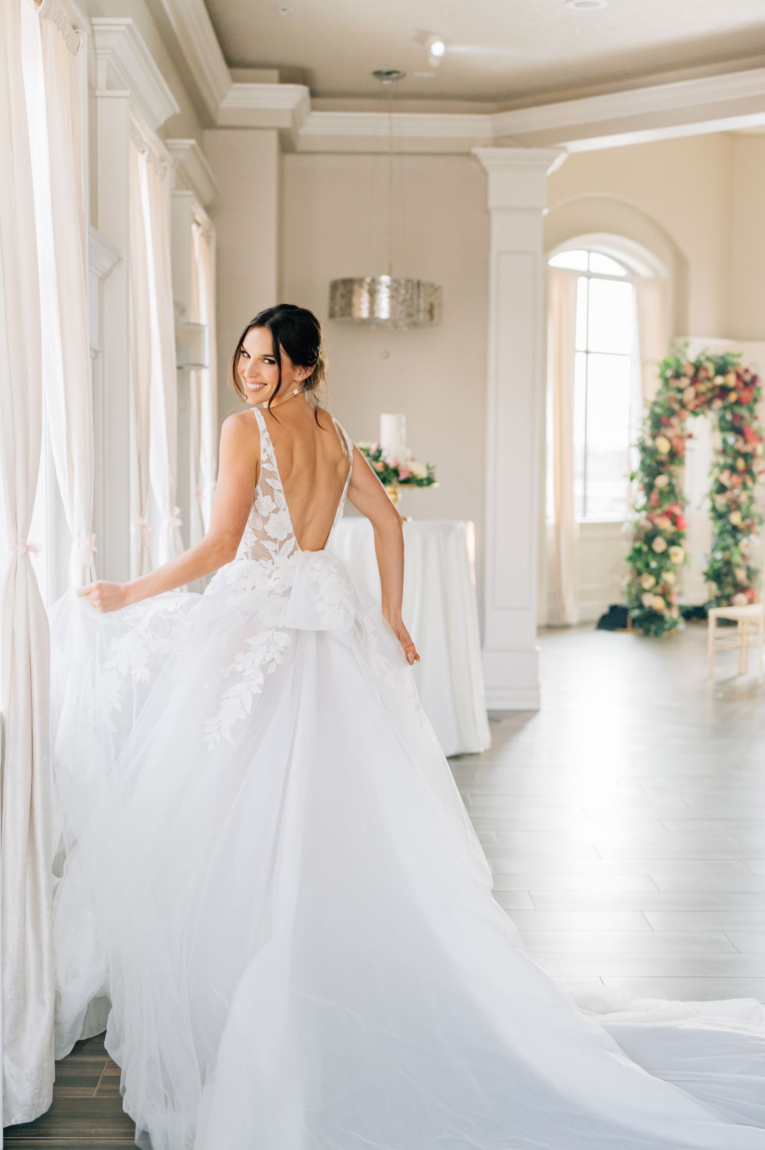 A bride wearing a white floral lace wedding dress sits thoughtfully by a window with her hand near her chin.
