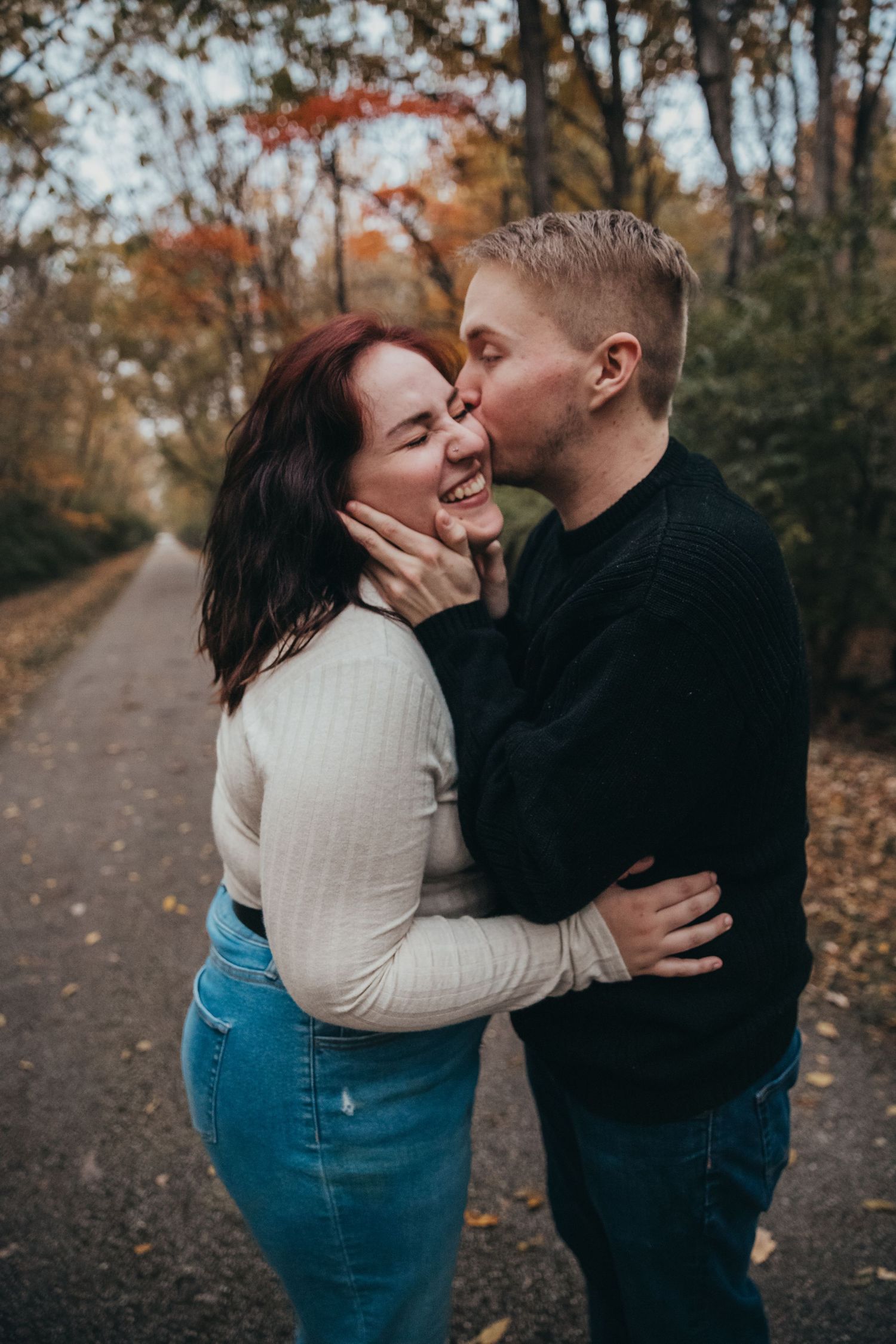 A romantic couple shares a playful moment on a path surrounded by autumn foliage.