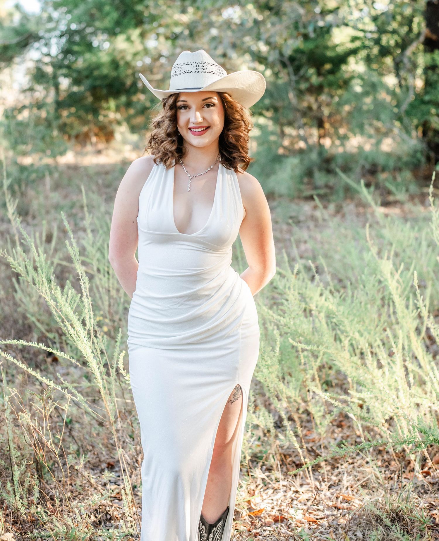 A person in a white dress and cowboy hat poses gracefully in a sunlit outdoor setting with natural greenery.