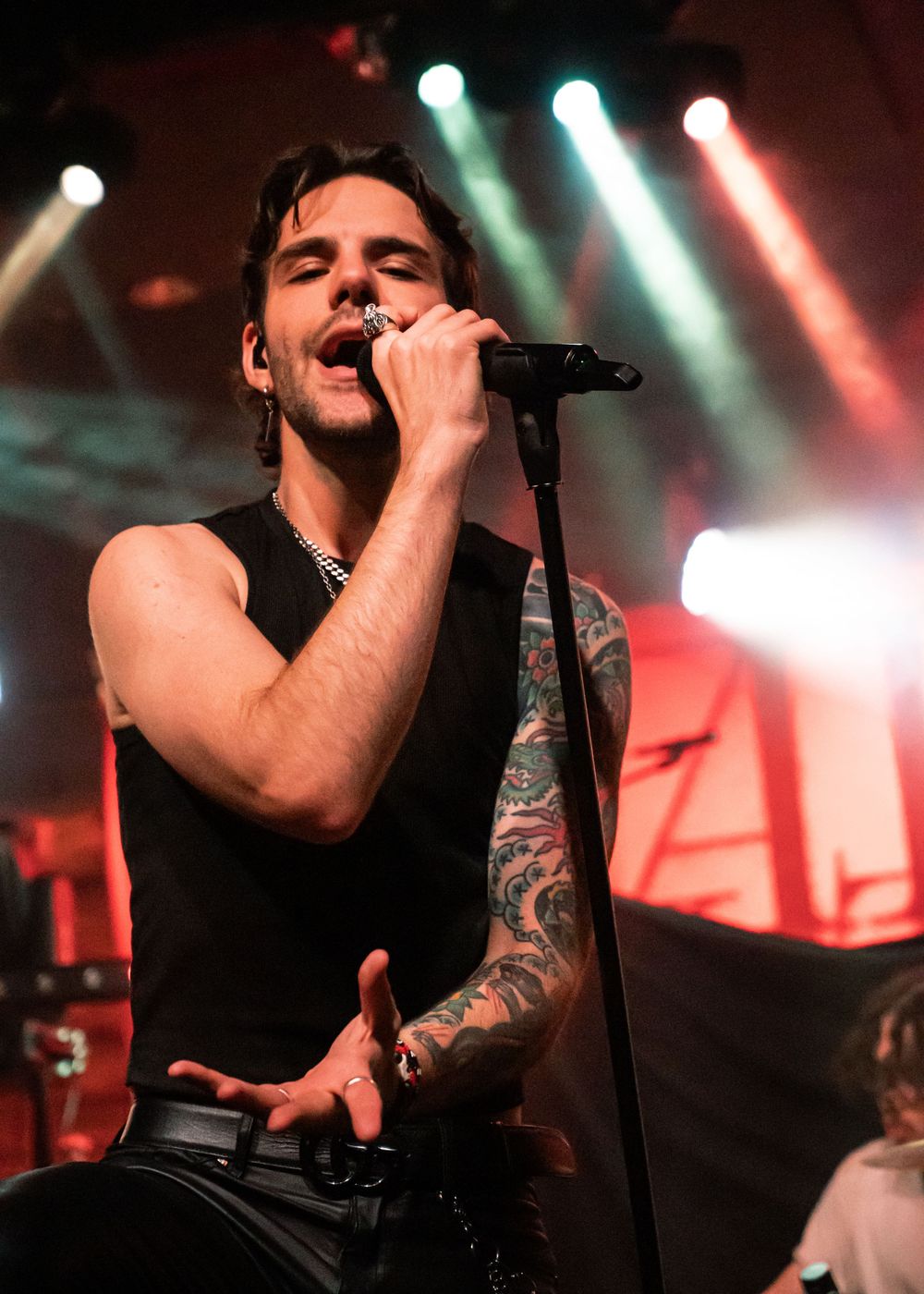 Tattooed male rock singer performing on stage with colorful stage lights, wearing a black sleeveless shirt and silver chain.