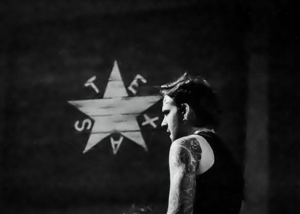 Black and white profile portrait of tattooed musician standing before star graffiti wall.