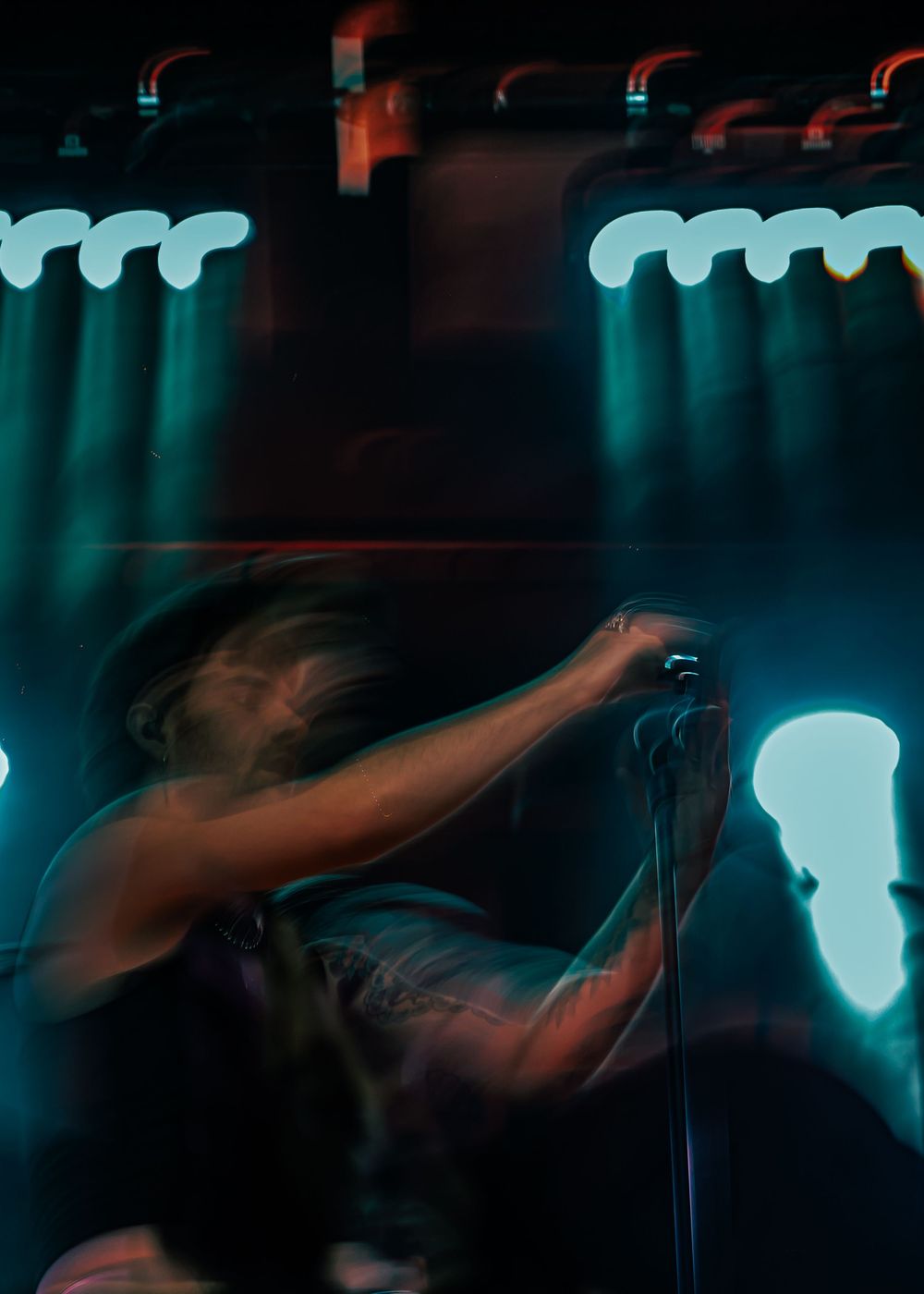 Blurred motion shot of energetic performer headbanging at microphone under teal stage lights.