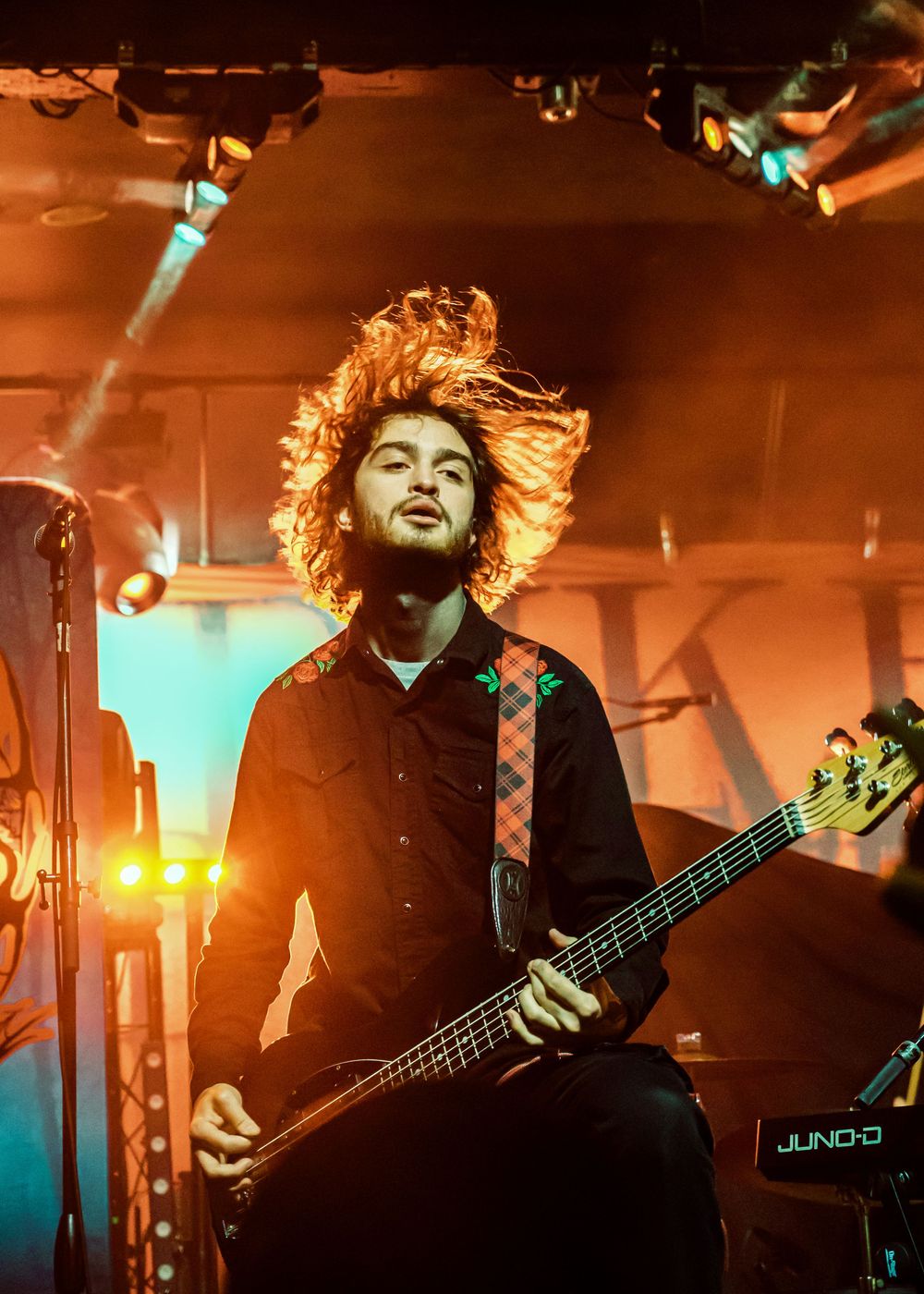 Curly-haired bassist playing guitar with hair flying under warm orange concert stage lights.