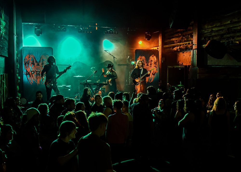 Packed crowd watching rock band perform on green and orange lit small venue stage.