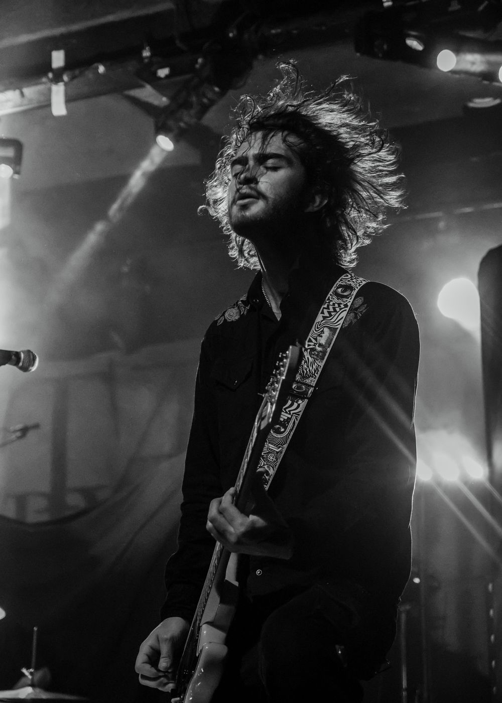 Black and white shot of long-haired guitarist headbanging with decorated guitar strap on stage.