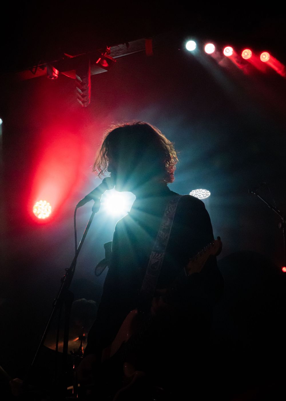 Silhouetted guitarist performing on stage with dramatic red and white spotlights beaming behind them at a live concert.