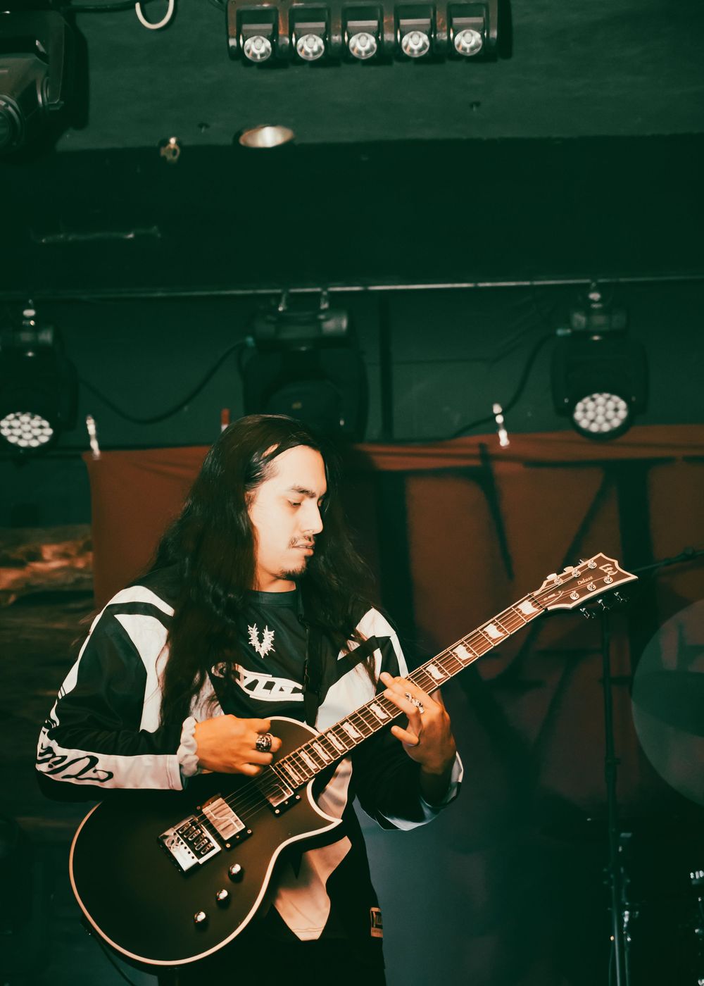 A long-haired musician plays a dark electric guitar on stage under colorful concert lights.