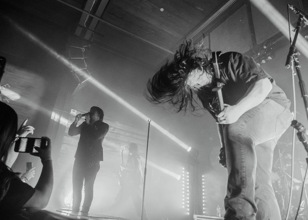 Black and white concert photo of rock musicians headbanging on stage with dramatic lighting beams.