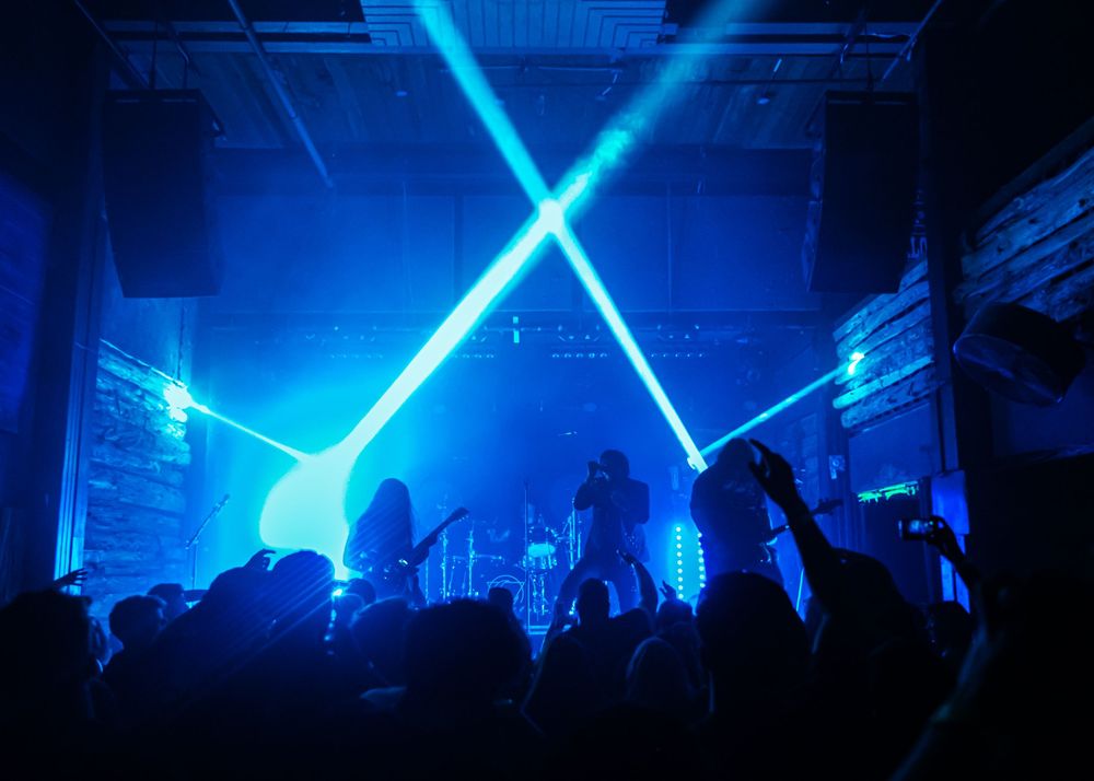 Concert crowd cheering with hands raised as blue laser lights cross above the stage in a dark music venue.