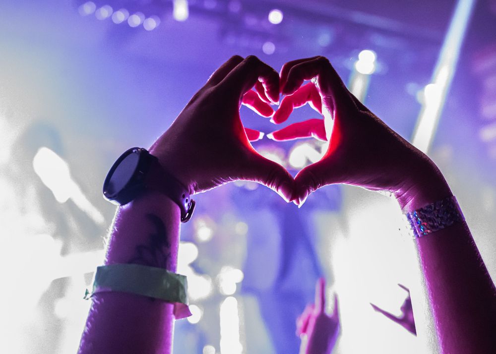 Two hands forming a heart shape at a concert with purple stage lighting glowing through.