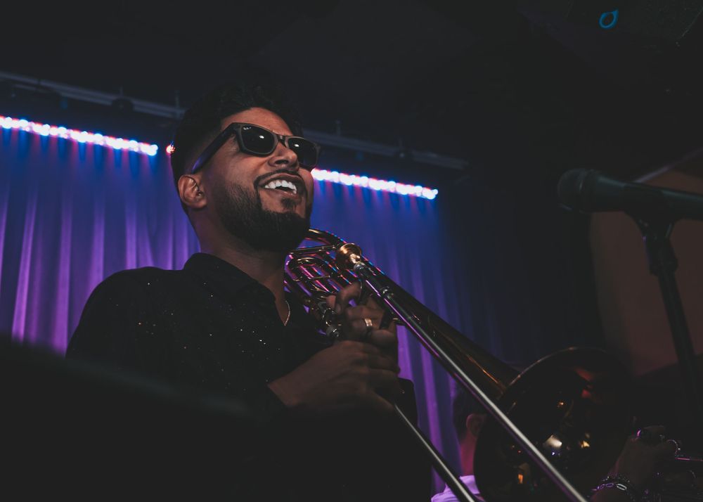 Smiling musician in sunglasses plays trombone on stage under purple spotlight at concert.