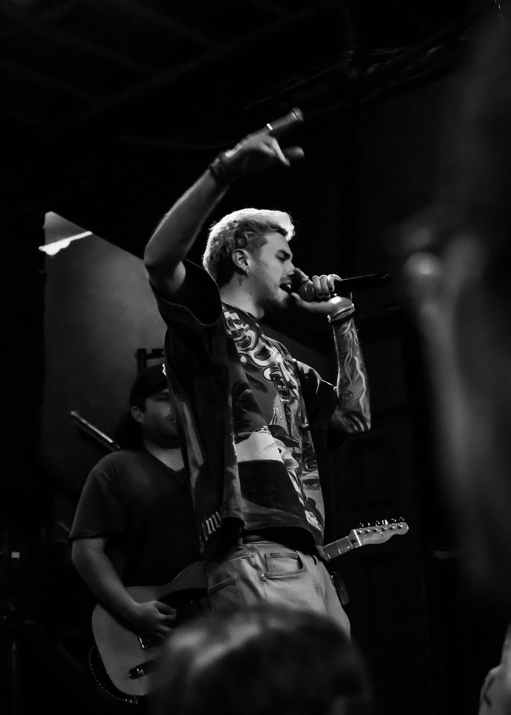 Black and white photo of a male vocalist singing on stage with hand raised, guitarist visible in background.