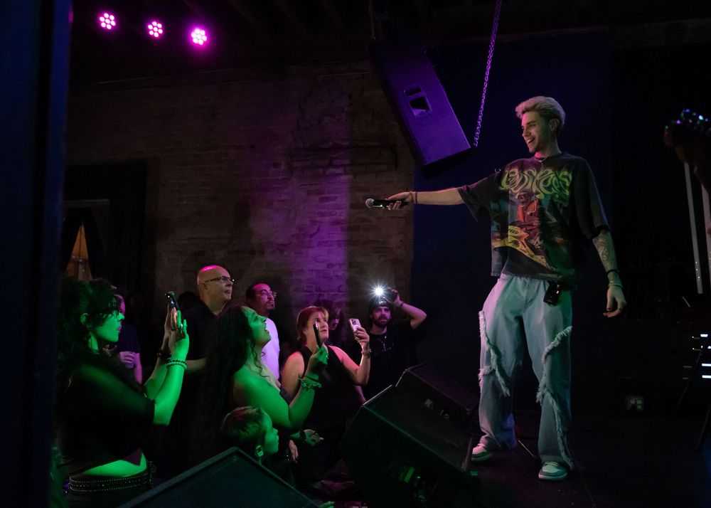 Performer in graphic tee pointing to crowd on a purple-lit stage during a live concert event.