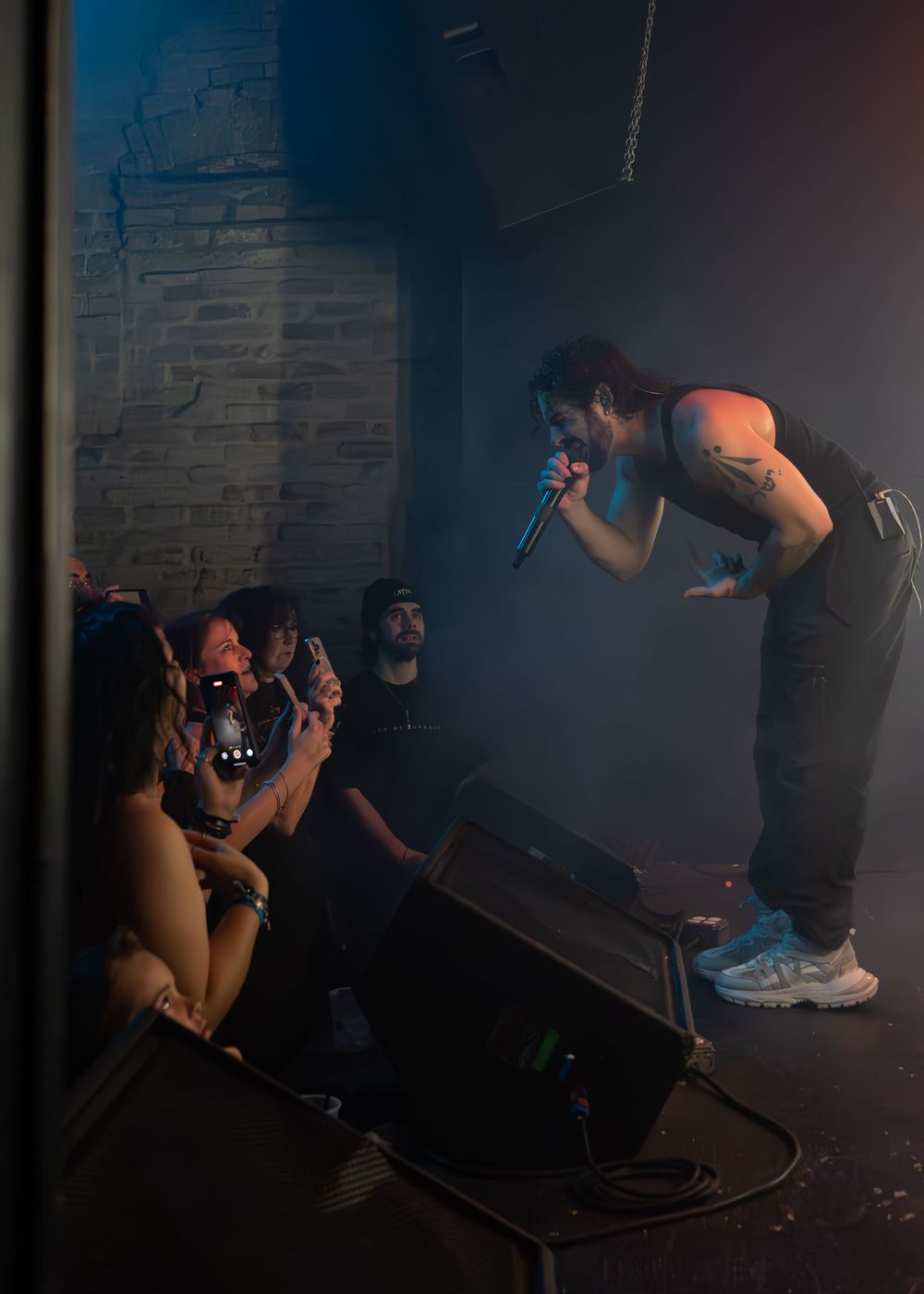 Rock vocalist leaning toward audience while performing on stage, fans photographing with phones below.
