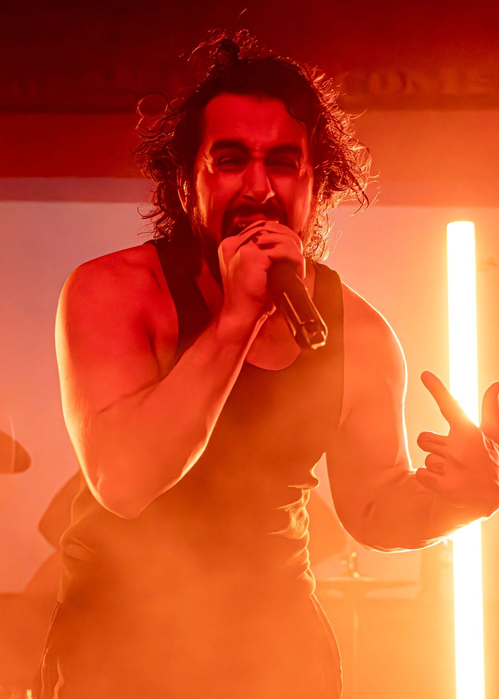 Male singer performing on stage in warm red light, holding microphone with curly hair and sleeveless top.