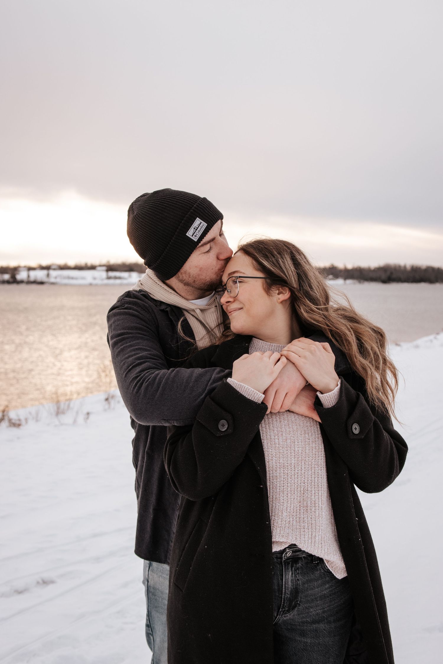 A couple embraces in a romantic winter moment by a snowy lakeshore at sunset.
