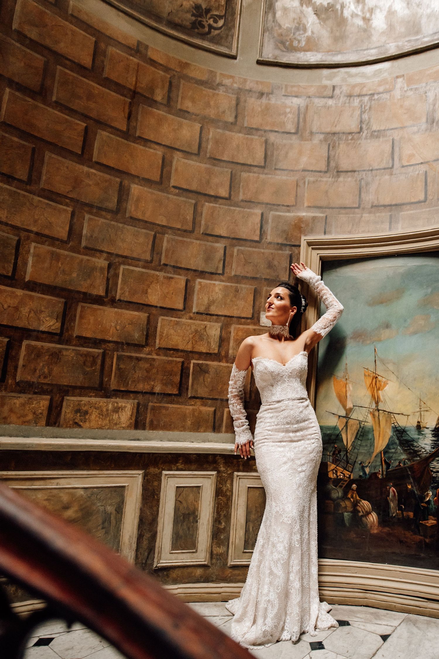 Elegant lace wedding dress photographed in a brick-walled room with vintage artwork.