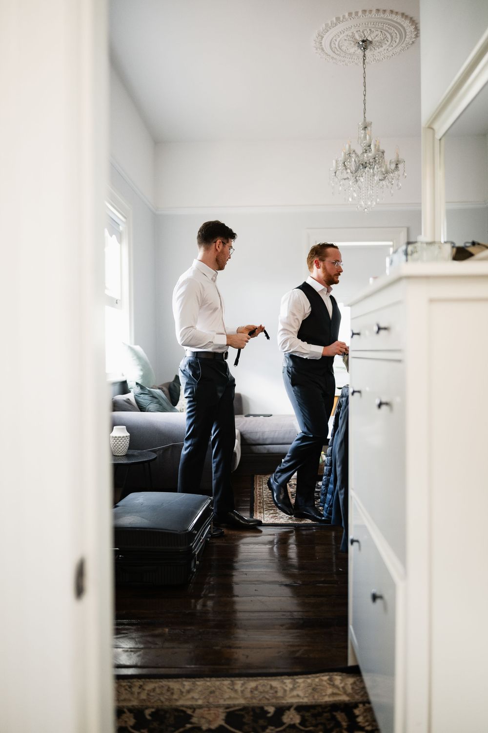 Two servers in formal attire prepare for service in an elegant white dining room with dark hardwood floors.