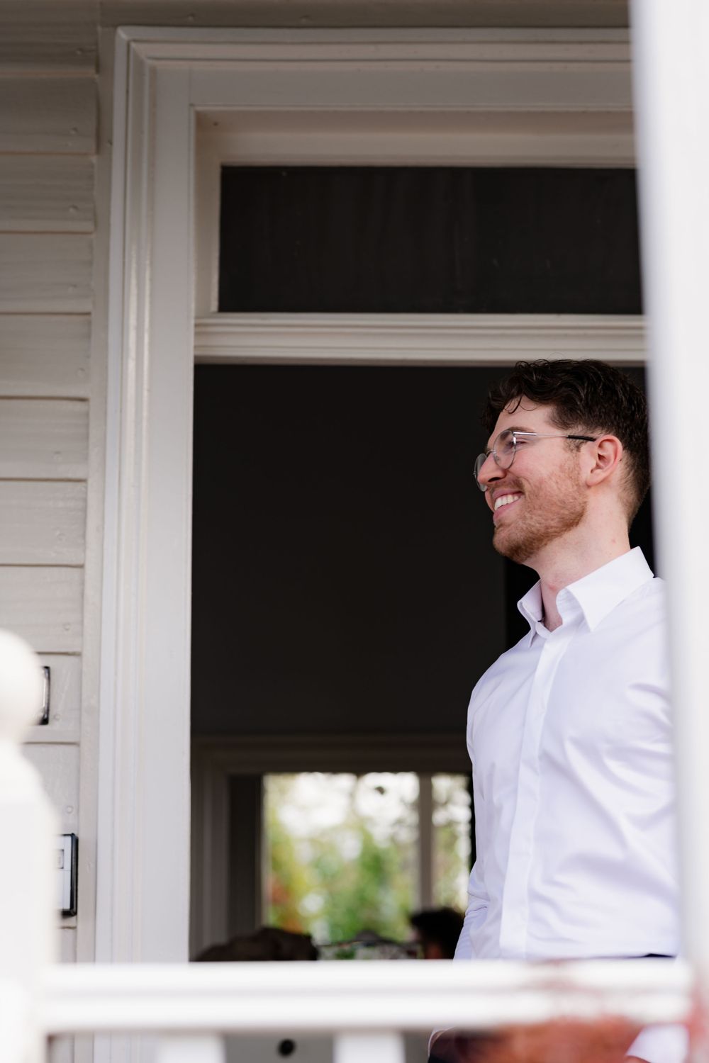A person in a white shirt smiles while sitting on a porch with white columns and railings.