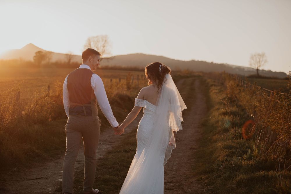 A bride and groom walk hand in hand along a rural path at golden hour sunset.