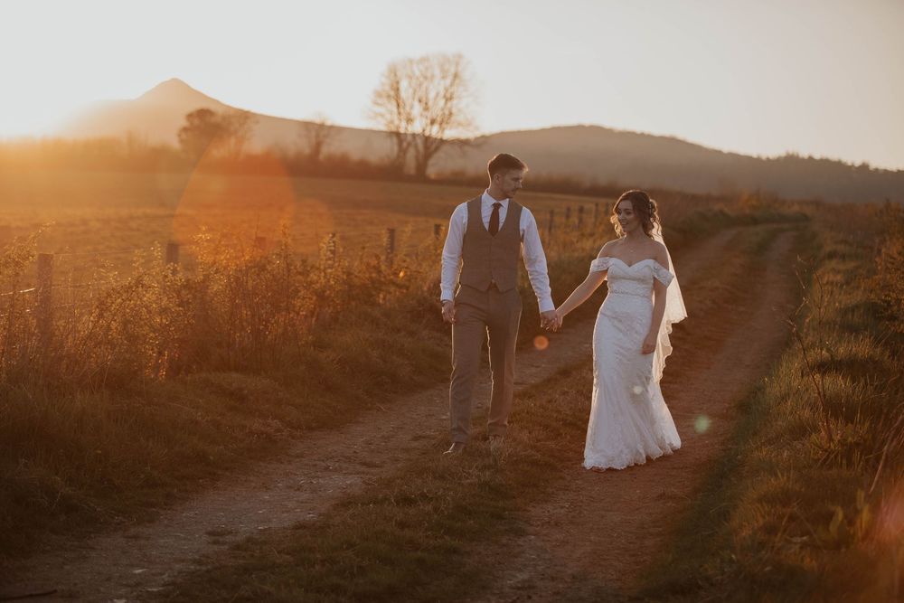 A bride and groom stroll down a dirt path surrounded by golden fields at sunset.
