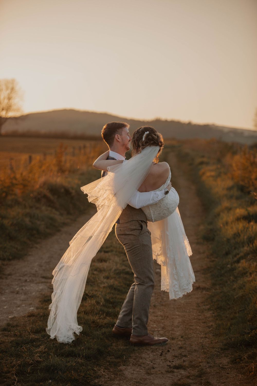 A groom lifts his bride on a scenic countryside path during a glowing sunset.