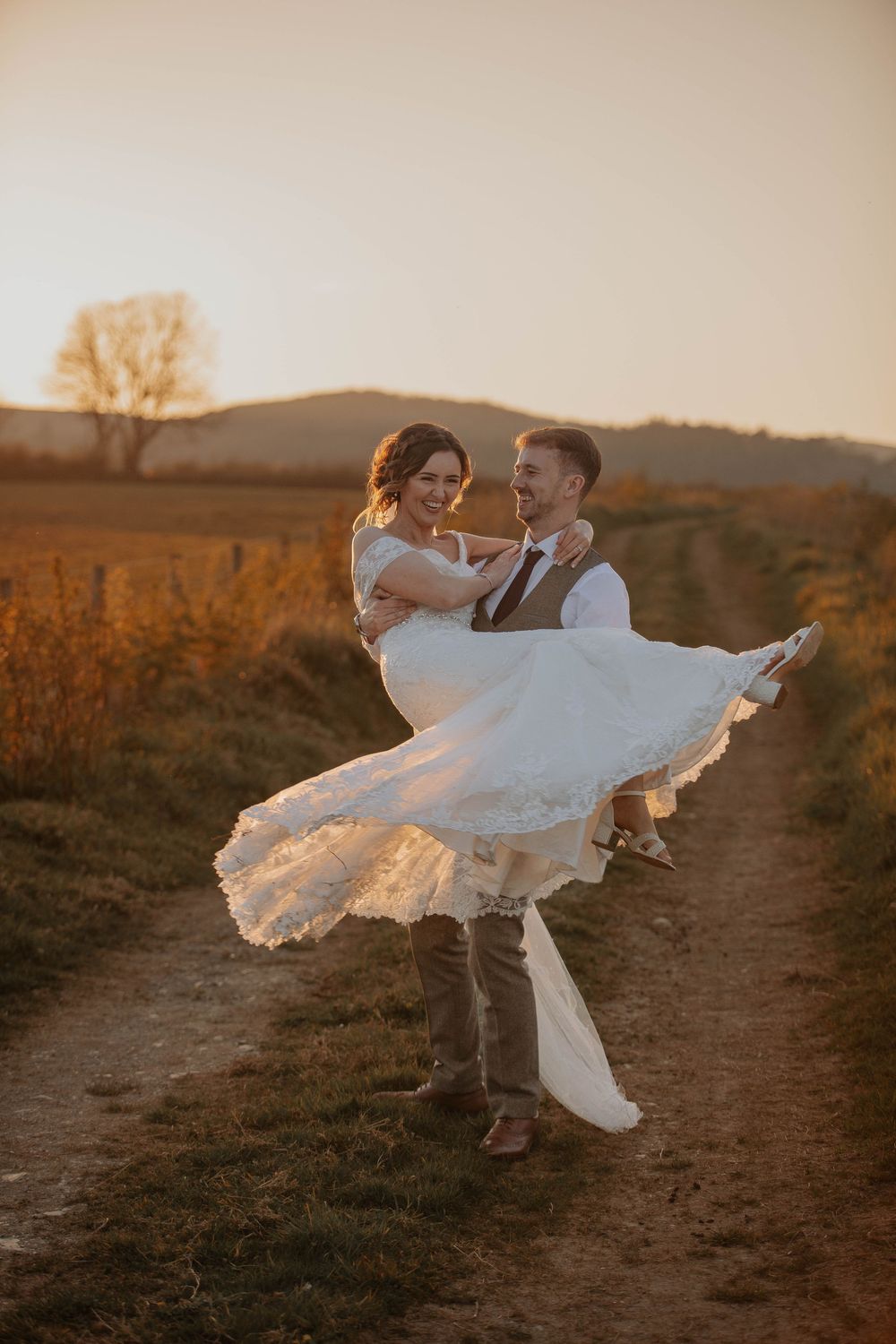 A groom spins his smiling bride in a white dress on a rural path at sunset.