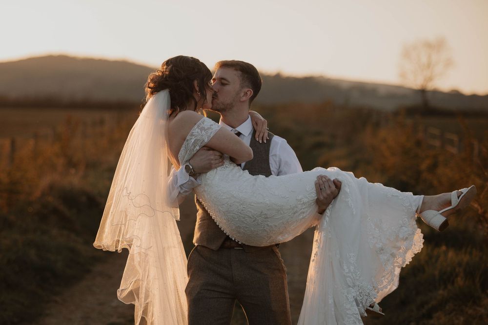 A groom carries his bride in his arms while kissing her in a golden sunset field.