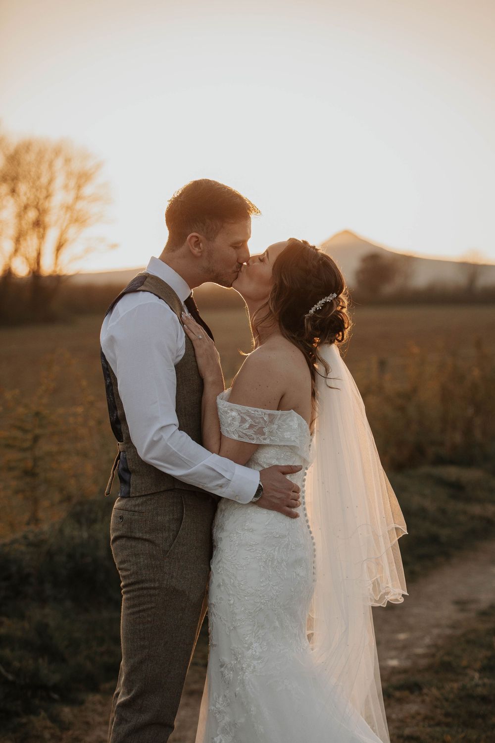 A bride and groom share a romantic kiss on a countryside path at golden hour.