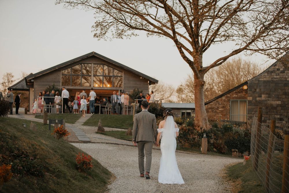A bride and groom walk toward a rustic barn wedding venue at dusk.