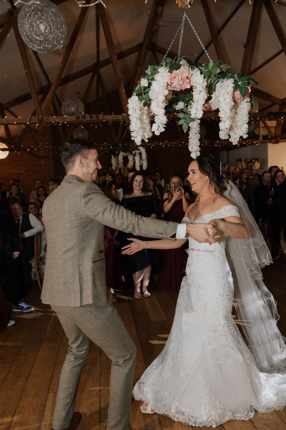 Bride and groom share a joyful first dance in a rustic barn wedding venue with floral chandelier.