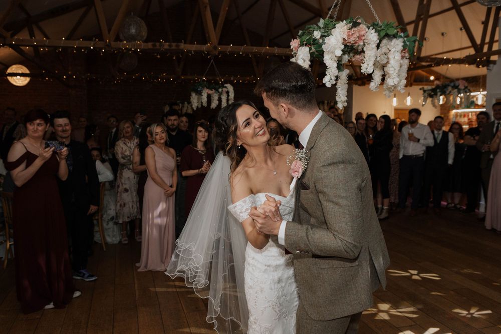 Newlyweds share an intimate slow dance moment surrounded by wedding guests in a warmly lit barn.