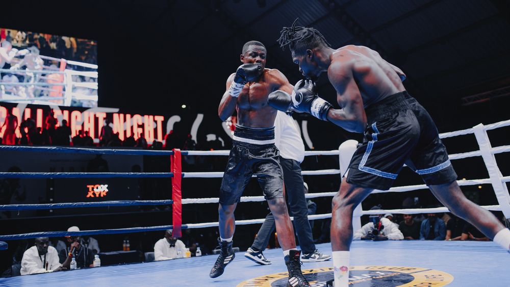Two boxers engage in an intense match inside a boxing ring with ropes and dark background.