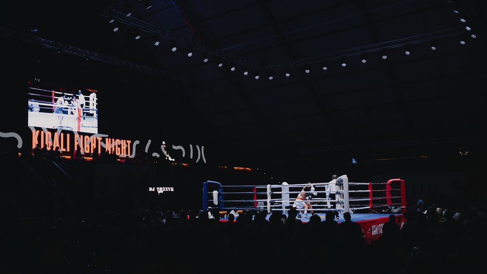Dark atmospheric boxing arena with illuminated ring and Kigali Fight Night signage visible.