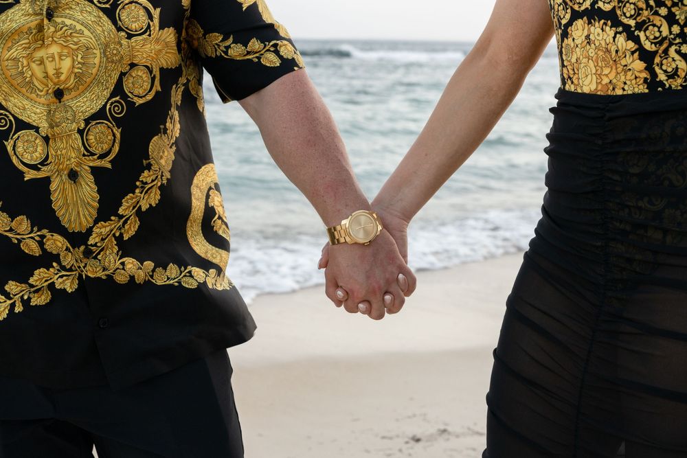 Couple in black and gold outfits holding hands while walking on a beach at sunset.