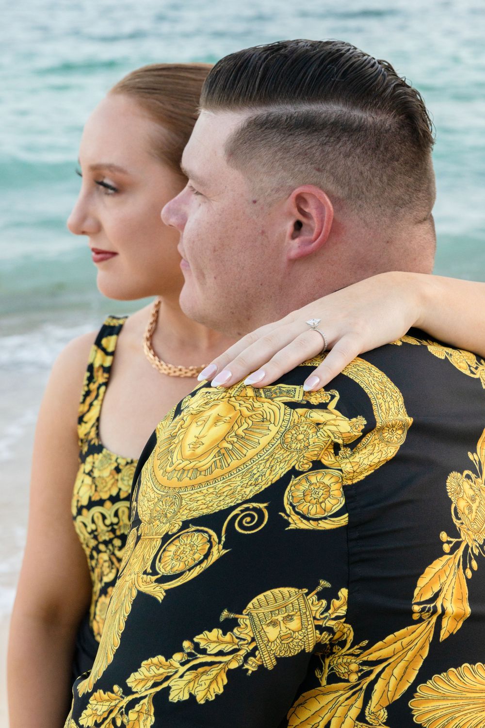A couple embraces by the ocean wearing coordinated black and gold baroque patterned formal attire.